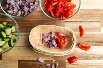 A wooden table with bowls of chopped vegetables including red onions, tomatoes, and cucumbers, alongside a stack of tortillas