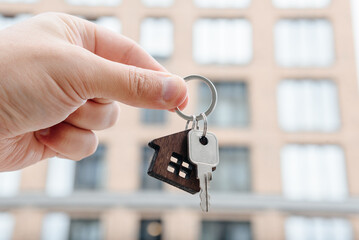 man hand with metal key, keychain in shape of private house, modern brown brick multi-storey apartment building in background, purchase of real estate, real estate services