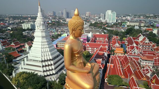 Aerial view of giant golden Buddha statue with white stupa and red temple roofs in Bangkok, Thailand. Traditional Buddhist architecture set against the modern city skyline.