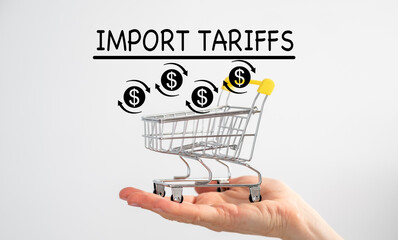 A hand holding an empty grocery basket and a sign on top with the text Import Tariffs and an upward...