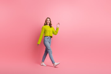 Cheerful young woman in trendy yellow sweater waving against pink background showcasing fun stylish...