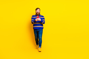 Fototapeta premium Cheerful bearded man wearing a colorful sweater poses confidently against a vibrant yellow background while using a smartphone.