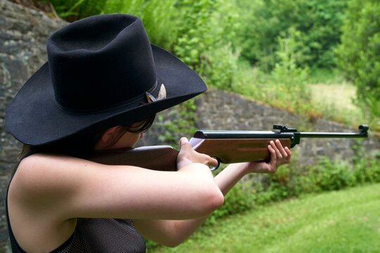 Woman in black hat aiming a rifle outdoors at a target range surrounded by greenery.