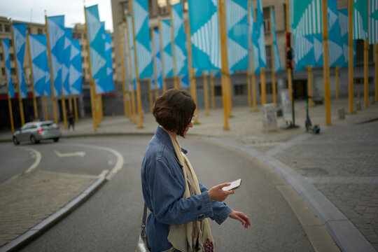 Woman on a street holding a phone with blue flags and buildings in the background. Brussels, Belgium