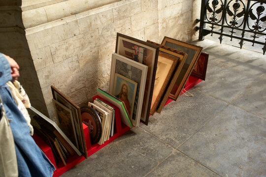 Assorted framed religious artworks and paintings displayed on a red stand against a stone wall. Brussels, Belgium
