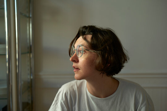 A young woman with glasses and short hair looks to the side in a softly lit room. Brussels, Belgium