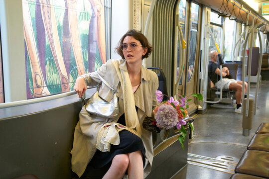 Stressed woman on a subway bench, wearing a trench coat, checking phone, holding bouquet of flowers. Brussels, Belgium