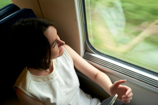 Woman in white top sitting by a train window holding a smartphone, gazing outside. Germany