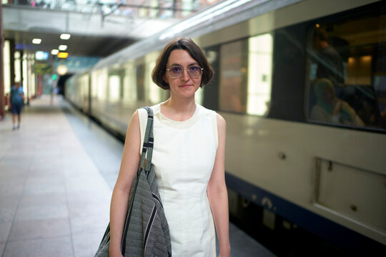 Woman in glasses and white dress at train station platform with a train in the background.