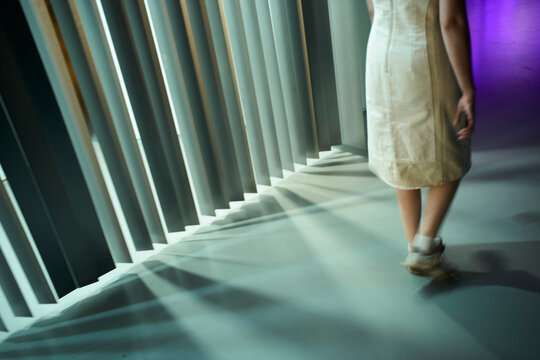 Woman in white dress walking past vertical blinds casting shadows on the floor. Germany