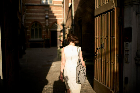 Woman in a sunlit alley with a bag, wearing a sleeveless white dress. Germany