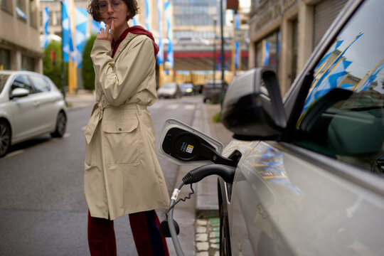 Woman in a trench coat stands by an electric car charging on a city street. Brussels, Belgium