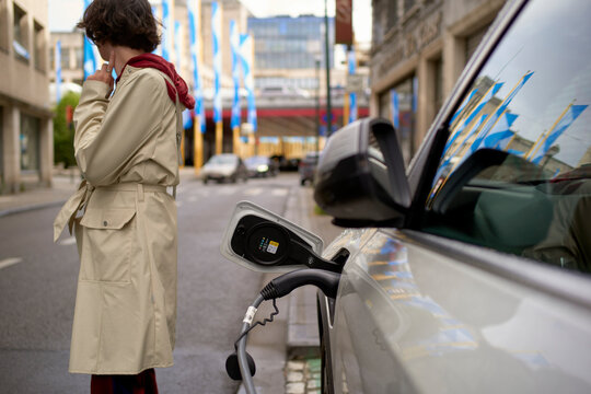 Person in beige coat standing near an electric car charging on urban street. Brussels, Belgium