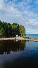 Fototapeta premium Enjoy the tranquility of Lake Saimaa, where two individuals explore the calm waters surrounded by verdant trees. An inviting van parked nearby enhances this serene escape in Finland.