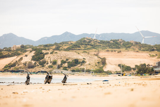 Three motorcycles parked on a sandy beach with distant hills and wind turbines in view. Beach near Phan Rang, Ninh Thuan, Vietnam
