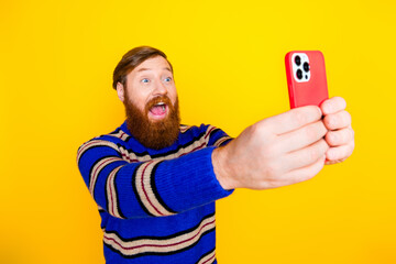 Excited bearded man taking a selfie with a smartphone against a bright yellow background wearing a...