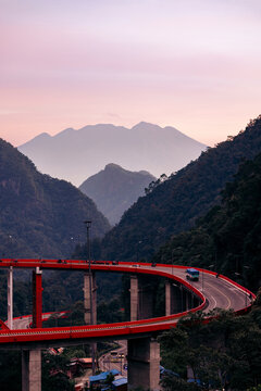 A winding red highway bridge curves through lush green mountains at sunset with a mountain backdrop.  Kelok 9, or the 9 hairpins, Harau, West Sumatra, Indonesia