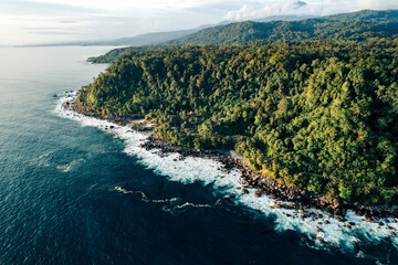 Aerial view of lush green forest coastline meeting the ocean under a cloudy blue sky. Krui coastline, Lampung, Indonesia