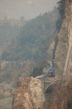 A narrow mountain road curves along steep cliffs with vehicles driving beside a river. Road H05, Chandi Bhanjyang, Nepal