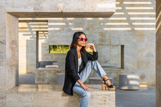 Stylish woman in sunglasses poses thoughtfully on a stone structure in a sunlit setting. Spain