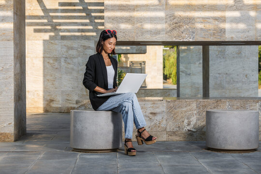 A woman sitting on stone ledge outside with a laptop, dressed casually in a blazer and jeans. Spain