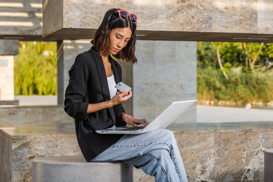 Woman in casual attire uses laptop and holds a credit card outdoors. Spain