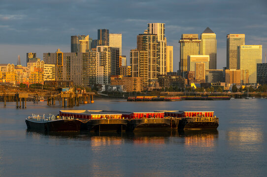 Golden sunset over a bustling city skyline with boats moored on a calm river. Canary Wharf, London, United Kingdom