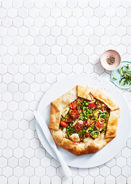 Rustic vegetable galette with tomatoes and peas on white hexagonal tile background.