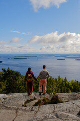 A couple stands hand in hand on a rocky cliff in Koli National Park, admiring the stunning view of calm waters and islands. The sky is bright and clear, creating a peaceful atmosphere.