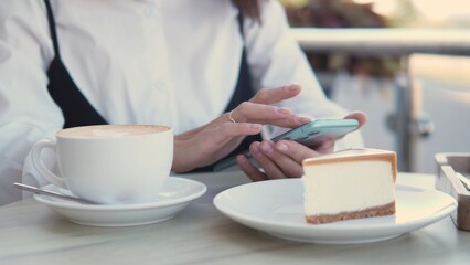 A woman sits at a table outdoors, using her smartphone. She has a cup of coffee and a slice of cheesecake nearby, engaging in a business discussion or personal work