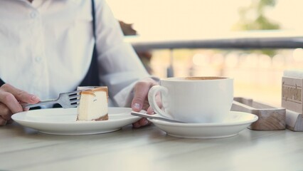A woman is seated at an outdoor cafe table, savoring a piece of cake while sipping coffee. It is a sunny afternoon, perfect for a relaxing treat