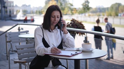 A woman in business attire sits at a cafe table, talking on her phone while taking notes. The setting is bright and daytime, with people walking in the background