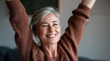 A joyful senior woman with gray hair smiles brightly while raising her arms in a relaxed indoor setting.