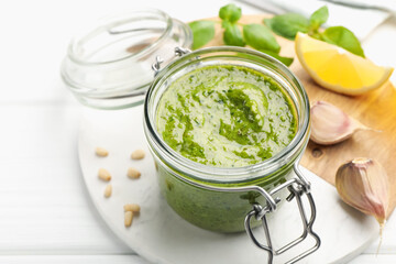 Tasty green pesto in glass jar and ingredients on white wooden table, closeup