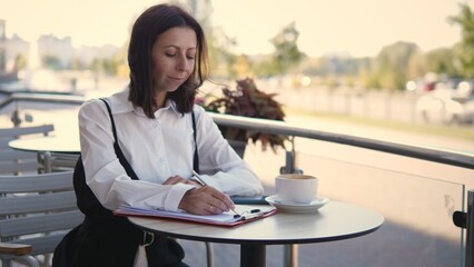 A woman sits at a cafe terrace, focused on writing in her notebook. She enjoys a cup of coffee while soaking in the lively atmosphere of the city during daytime