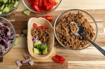 A bowl of cooked ground meat next to a taco shell filled with diced vegetables, including cucumbers, tomatoes, and onions, with additional chopped vegetables in bowls around it