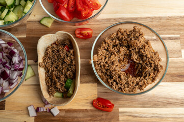 A bowl of ground meat next to a tortilla filled with chopped vegetables, surrounded by bowls of diced tomatoes, cucumbers, and onions on a wooden table