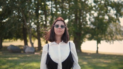 A confident woman stands outdoors in a sunny setting, wearing sunglasses and a stylish outfit. She appears relaxed while enjoying a moment in nature