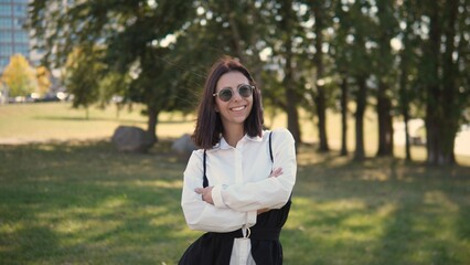 A confident woman stands with crossed arms in a park surrounded by trees. She wears sunglasses and a stylish outfit, smiling as she enjoys a sunny day in the city outdoors