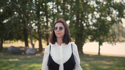 Obraz premium A confident business woman stands in a park on a sunny afternoon, wearing sunglasses and a stylish outfit. She appears relaxed and thoughtful, enjoying the tranquil atmosphere around her