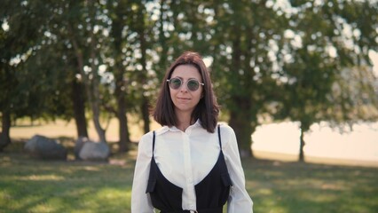 A business woman stands outdoors in a park, wearing a stylish outfit and sunglasses. She smiles confidently as sunlight filters through the trees around her