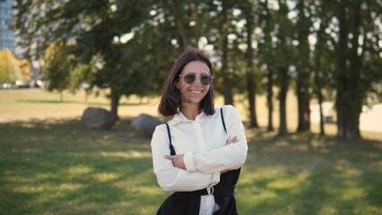 A businesswoman stands confidently in a park, smiling with her arms crossed. She is wearing sunglasses and a stylish outfit. The sun shines brightly, showcasing a vibrant green landscape