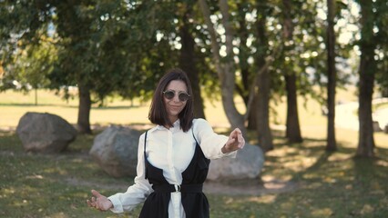 A business woman stands confidently in a park wearing sunglasses and a stylish outfit. She is gesturing as if explaining something important while surrounded by trees and rocks