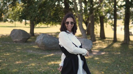 A business woman stands in a park, smiling and wearing sunglasses. She is dressed in a stylish outfit, enjoying the fresh air and greenery around her during a sunny day