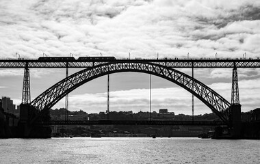 Obraz premium Silhouette of the iconic bridge from Oporto to Vila Nova de Gaia over the Douro River in Portugal. A modern metro train and numerous pedestrians cross the double bridge against the backdrop of the sun