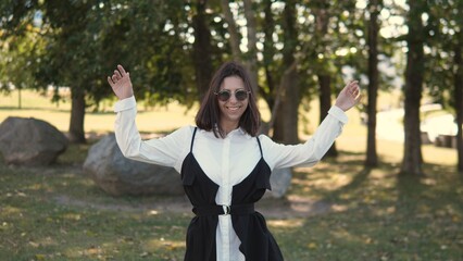 A business woman smiles happily while standing in a park on a sunny day. She wears a chic outfit with stylish sunglasses and is surrounded by greenery and rocks, creating a lively atmosphere