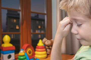 A sleepy boy rubs his eyes in a room with toys under window
