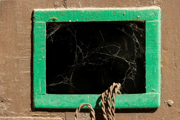 Close-up view of a small, rustic window in a door with cobwebs inside and iron wire hanging outside.