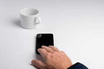 A hand reaching for a smartphone on a white surface, with a white cup in the background