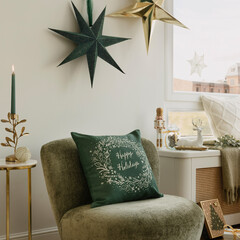 Green velvet chair with “Happy Holidays” cushion under paper stars, candle, nutcracker and reindeer on the windowsill create a warm, modern Christmas corner.